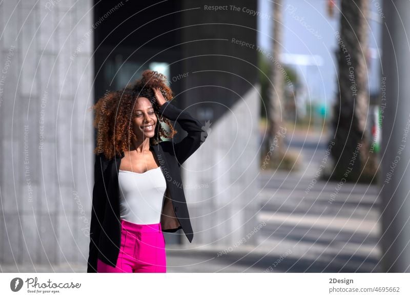 Front view of a young beautiful curly woman wearing elegant clothes and handbag while standing in the street in sunny day front view afro american outdoors