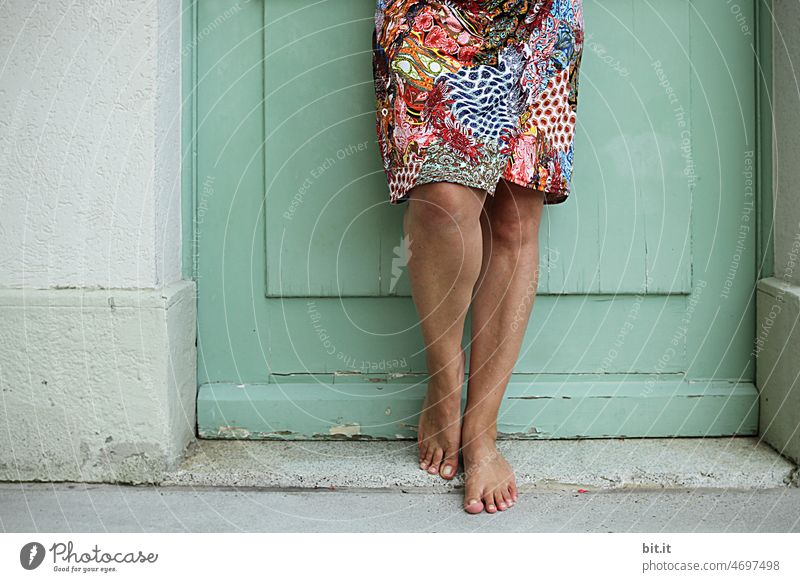 Paisley - pattern - dress in colorful, over brown tanned slender legs - in front of door made of wood in mint - and gray concrete wall in summer heat. Legs