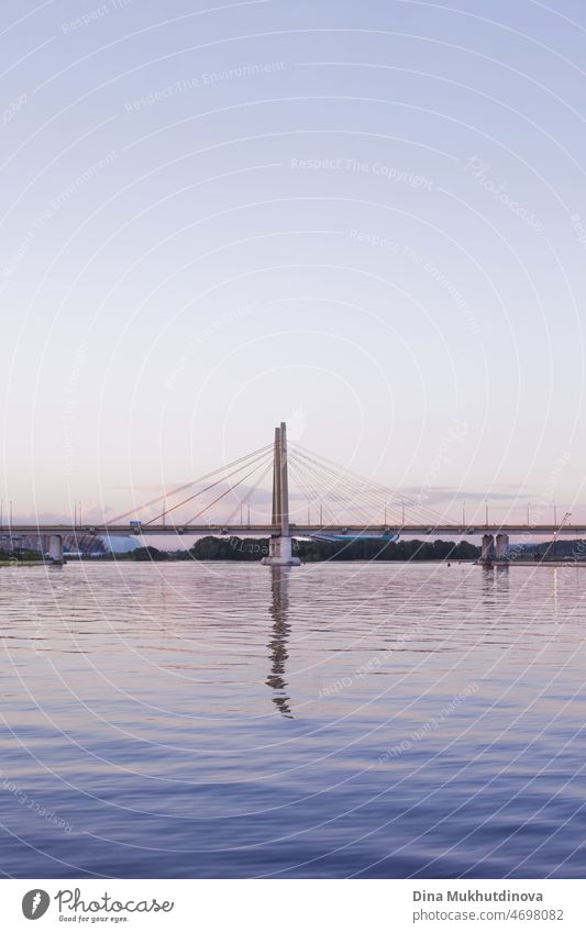 Suspension bridge at sunset at blue hour. Cloudless sky, bridge reflected in river water. Urban cityscape and modern architecture. Vertical urban summer background.