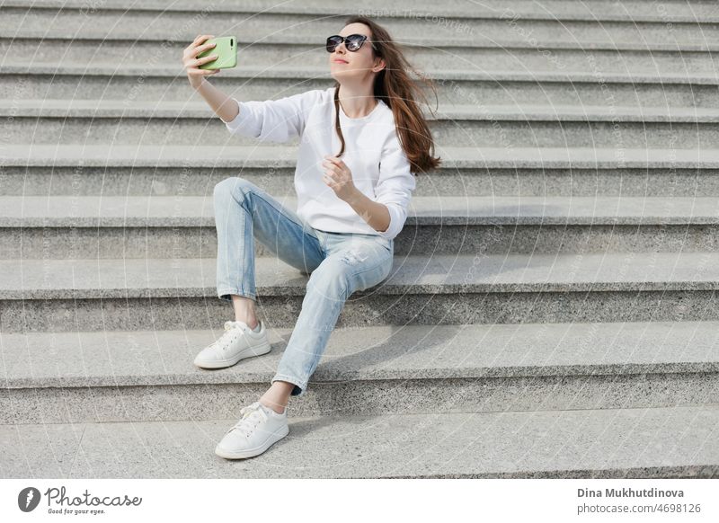 Young millennial woman in white shirt taking a selfie or videochatting on mobile phone sitting on the steps of a building. Female student using technology sitting on staircase steps of university campus. Real people with mobile phones.