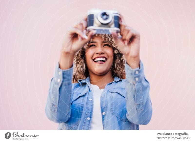 beautiful happy hispanic woman with afro hair holding vintage camera. pink background photographer city touch smiling spring casual clothing lifestyle