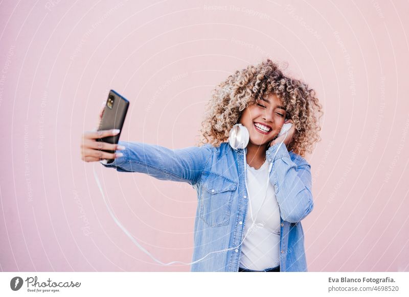 portrait of smiling hispanic woman with afro hair in city using mobile phone and headset. lifestyle selfie technology internet wireless picture device music