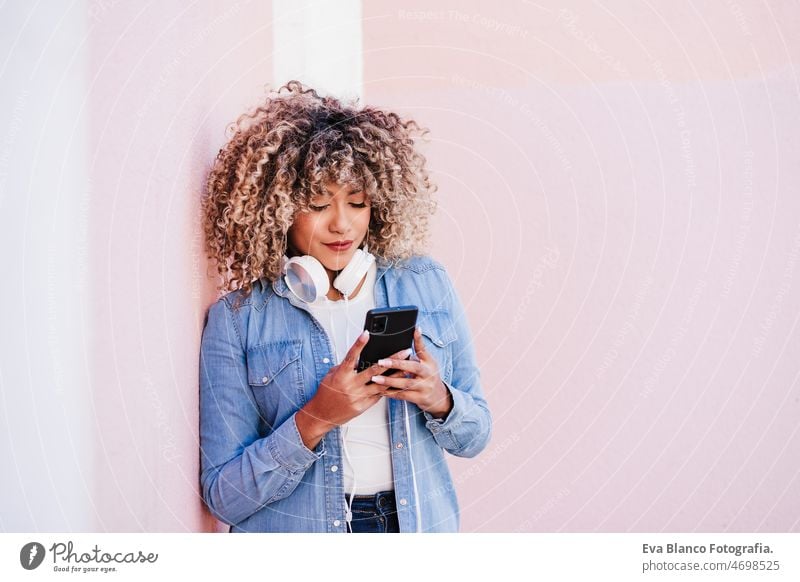 portrait of smiling hispanic woman with afro hair in city using mobile phone and headset. lifestyle selfie technology internet wireless picture device music