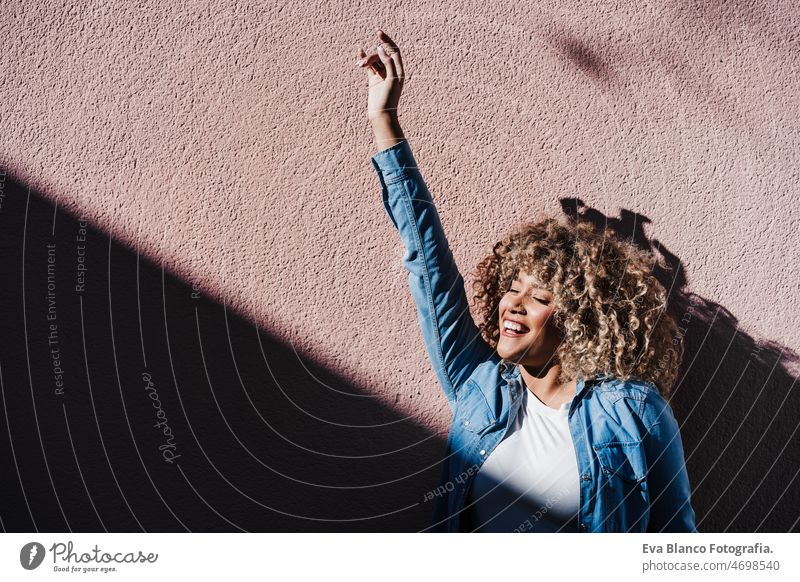 smiling afro hispanic woman in sunny day with eyes closed enjoying outdoors. City lifestyle,spring happy city pink casual clothing curly hair urban adult