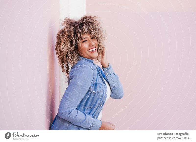 portrait of smiling hispanic woman with afro hair in city during spring. Urban lifestyle pink happy casual clothing curly hair outdoors urban adult laughing