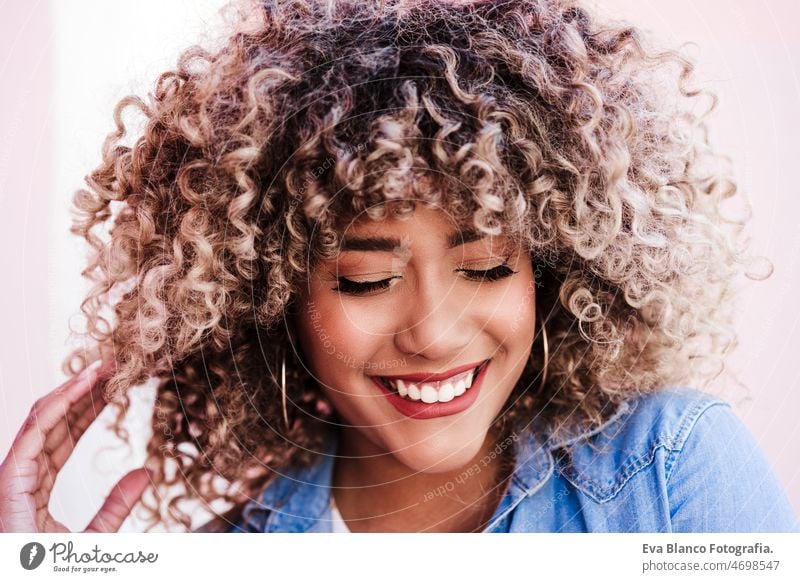 portrait of beautiful hispanic woman, afro hair outdoors in spring.City pink background. lifestyle happy smiling city eyes closed casual clothing curly hair