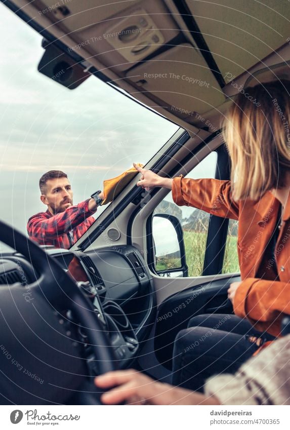 man cleaning motorhome windshield while woman pointing stain perfectionist rinsing glass cloth view from inside through the glass helping concentrated outdoors