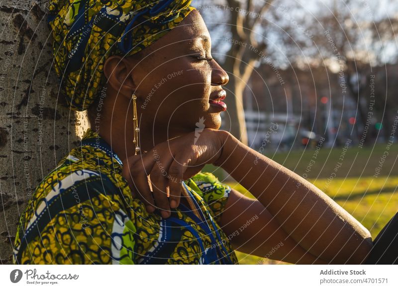 African woman resting near tree park weekend summer city tradition trunk daytime female street ethnic african american black portrait sit headscarf headwear