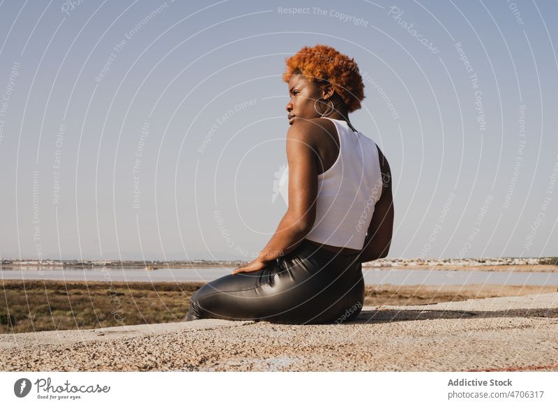 Black woman sitting on shore of sea in sunny day rest trendy style sand summer thoughtful individuality pants tranquil relax leather nature idyllic slim coast