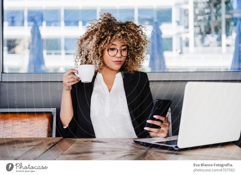 smiling hispanic afro business woman in cafe working on laptop and mobile phone. tech and lifestyle computer networking drinking coffee terrace outdoors goggles