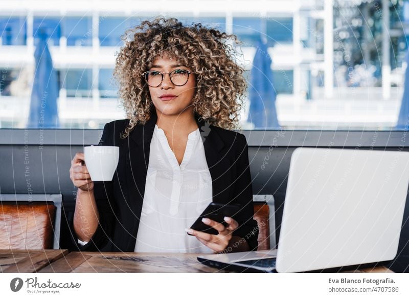 confident hispanic afro business woman in cafe working on laptop and mobile phone.tech and lifestyle computer networking drinking coffee terrace outdoors