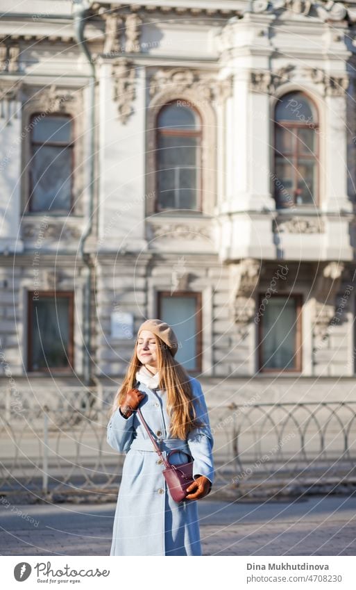 Young beautiful woman in a light blue coat standing on the street near old building in the city in sunlight on sunny day. Candid lifestyle portrait of a woman, smiling.