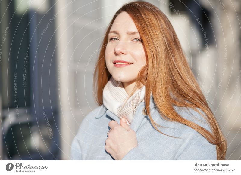 Young beautiful woman in a light blue coat standing on the street in the city in sunlight on sunny day. Candid lifestyle portrait of a woman, looking at the camera and smiling.