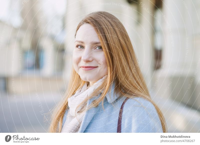 Young beautiful woman in a light blue coat standing on the street in the city. Candid lifestyle portrait of a woman, looking at the camera and smiling. happy