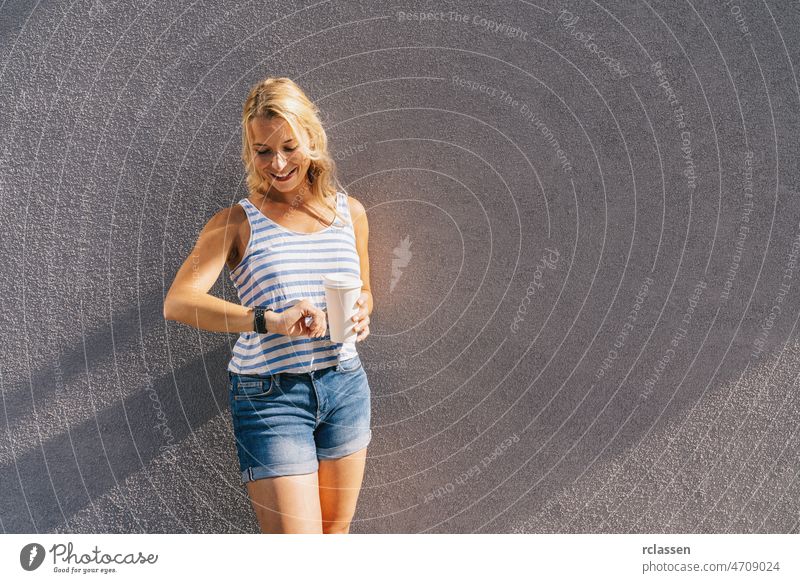 Stylish happy young woman looking to her clock. She holds paper coffee to go and enjoying the sunlight in the city. portrait of smiling girl , lifestyle concept image.