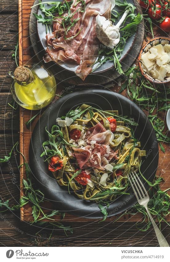 Green tagliatelle pasta with ham, parmesan cheese, arugula and tomatoes on black plate green rustic wooden kitchen table ingredients olive oil cutlery top view