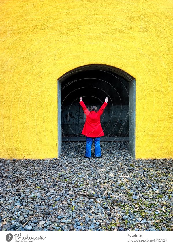 Color Contest | Red Coat II - She loves colors. In a bright yellow archway, with her red coat and blue denim pants she stands out super against the ground with gray pebbles.