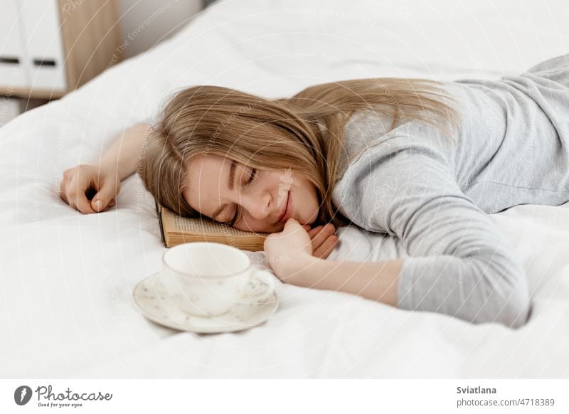 A young girl lies with her eyes closed, holding a book in her hands, lying on the bed at home woman reading bedroom morning rest house grey beautiful coziness