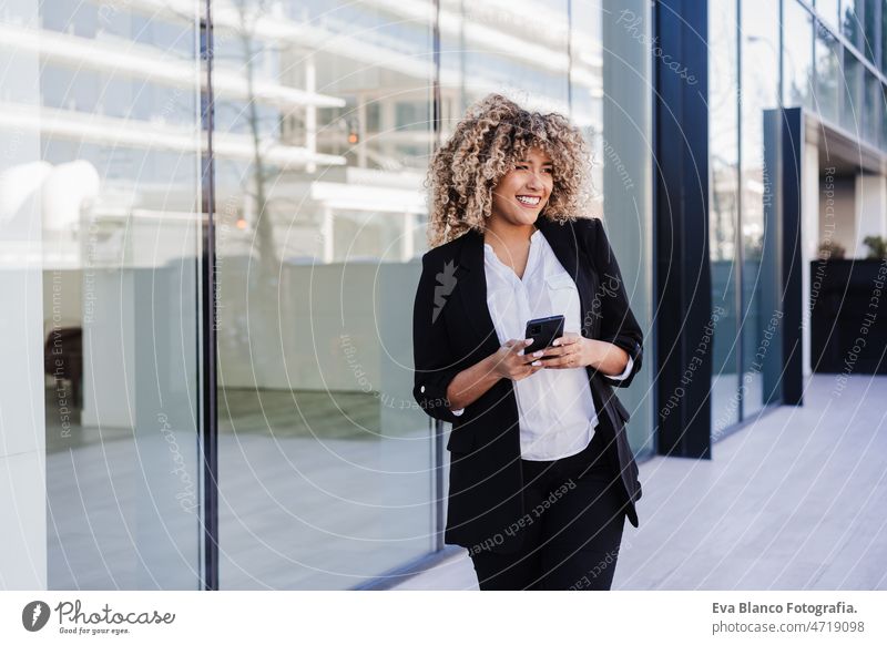 beautiful smiling business woman using mobile phone and headphones in city. Buildings background afro hispanic skyscraper building young curly hair music