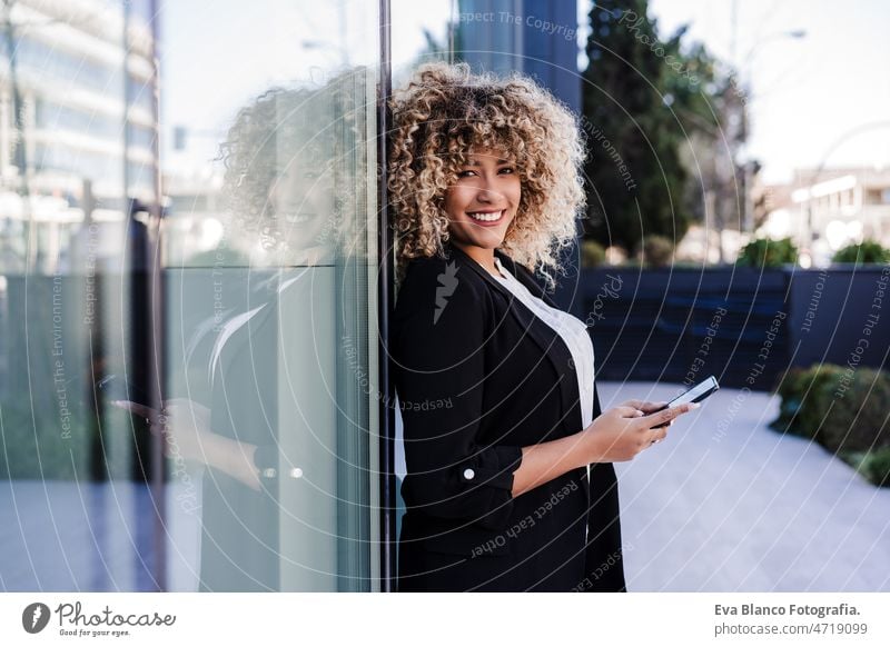 beautiful smiling business woman using mobile phone and headphones in city. Buildings background afro hispanic skyscraper building young curly hair music