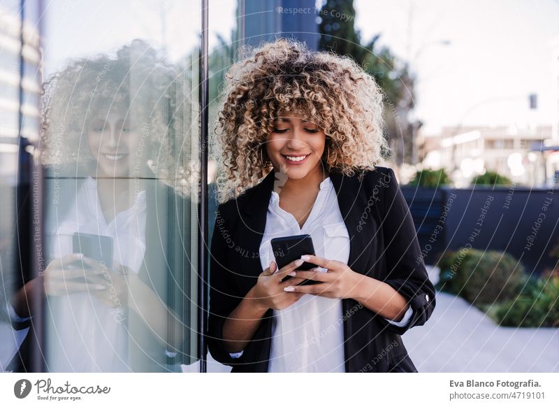 beautiful smiling business woman using mobile phone and headphones in city. Buildings background afro hispanic skyscraper building young curly hair music