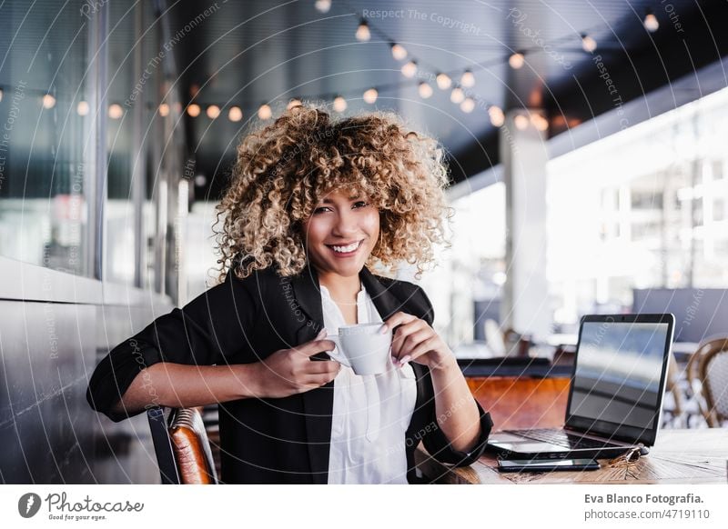 confident hispanic business woman in cafe working on laptop and mobile phone. tech and lifestyle computer afro networking drinking coffee terrace outdoors