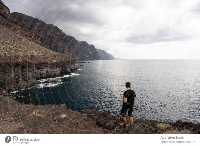 Traveler standing on rocky coast of rippling ocean man tourist freedom admire vacation mountain seascape gloomy wanderlust trip male photo camera traveler shore