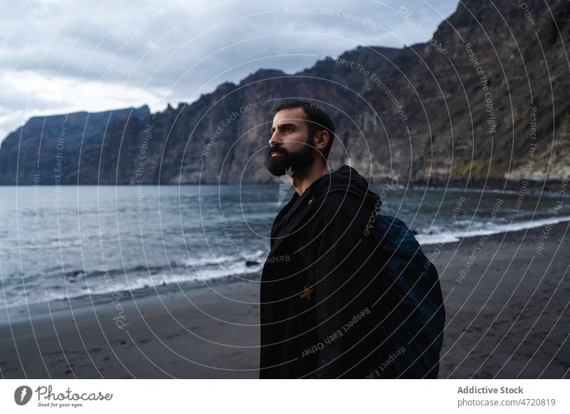 Serious man with backpack standing on sandy beach traveler admire cliff sea rock coast explore journey male nature slope tourist steep rocky ocean trip shore