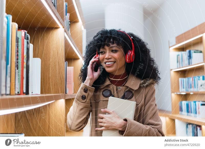 Positive black female meloman speaking on smartphone in library woman using shelf book headphones conversation bookcase positive phone call student bookshelf