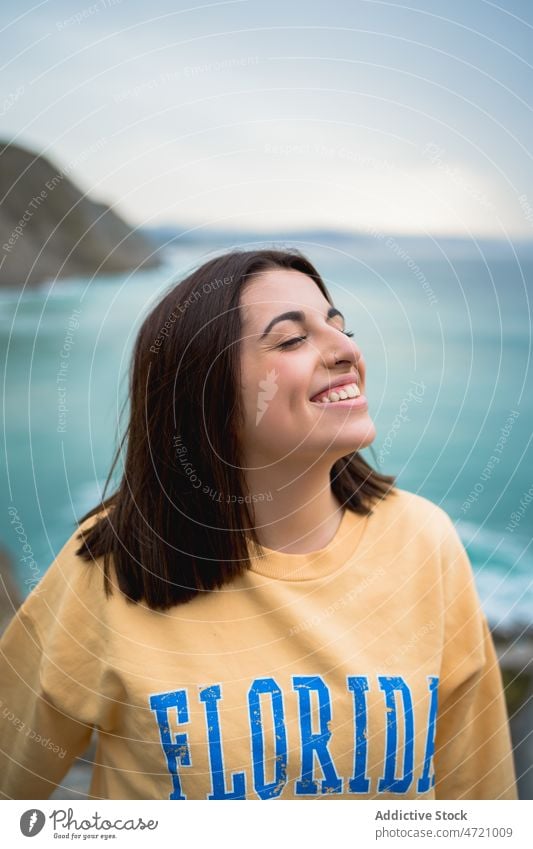 Cheerful woman standing on rocky seashore coast nature pastime enjoy trip leisure adventure travel seaside feminine admire eyes closed pleasure recreation smile