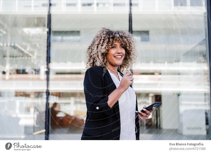 beautiful smiling business woman using mobile phone and headphones in city. Buildings background afro hispanic skyscraper building young curly hair music