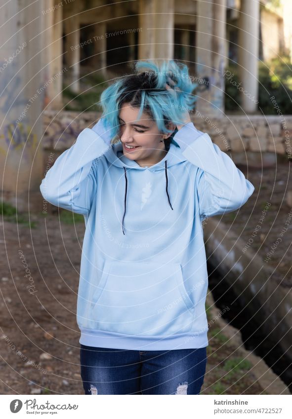 Blue haired Smiling Teenage girl in light blue oversize hoodie stays under bridge Teenager mockup jeans blue haired teen girl outdoors bridge pillars graffiti