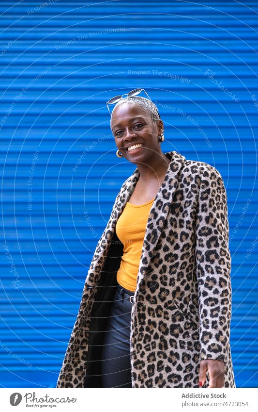 Positive black woman near blue wall street style appearance outfit trendy city urban garment smile african american female glad content happy positive coat