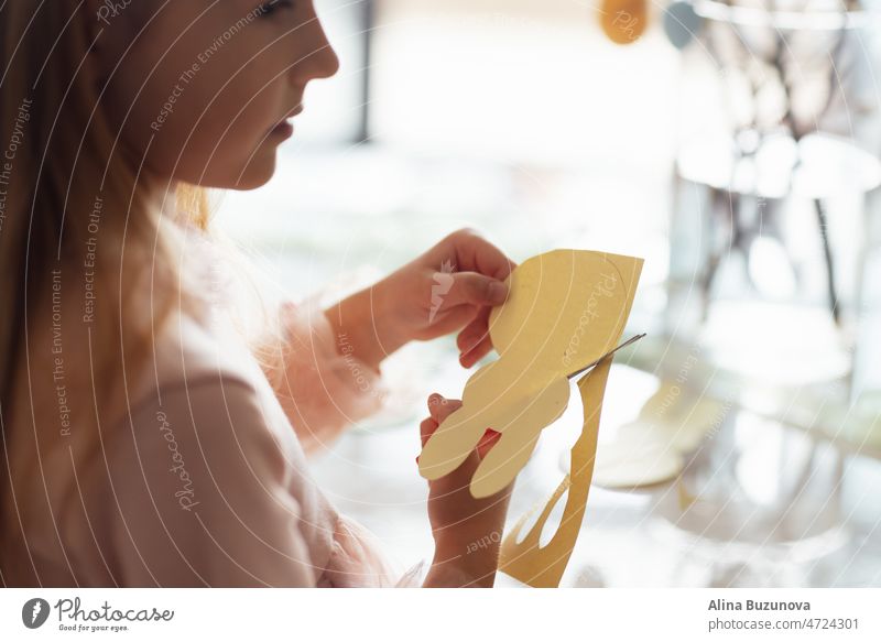 happy caucasian toddler girl eight years old at home in living room making garland with paper bunny. Stay home during Coronavirus covid-19 pandemic easter egg