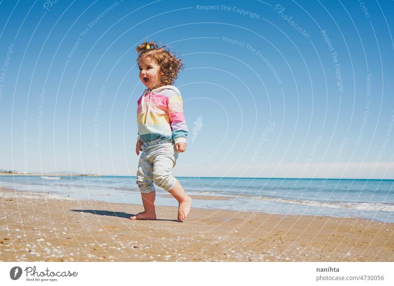 Wide view of a little girl wearing rainbow hoodie playing at the beach freedom happiness holidays baby toddler family sea shore playful real lifestyle people