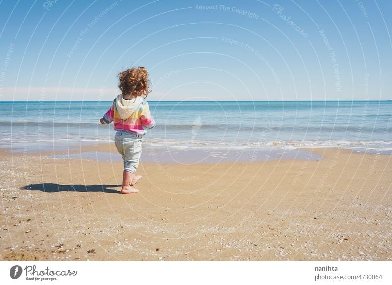 Wide view of a little girl wearing rainbow hoodie playing at the beach freedom happiness holidays baby toddler family sea shore playful real lifestyle people