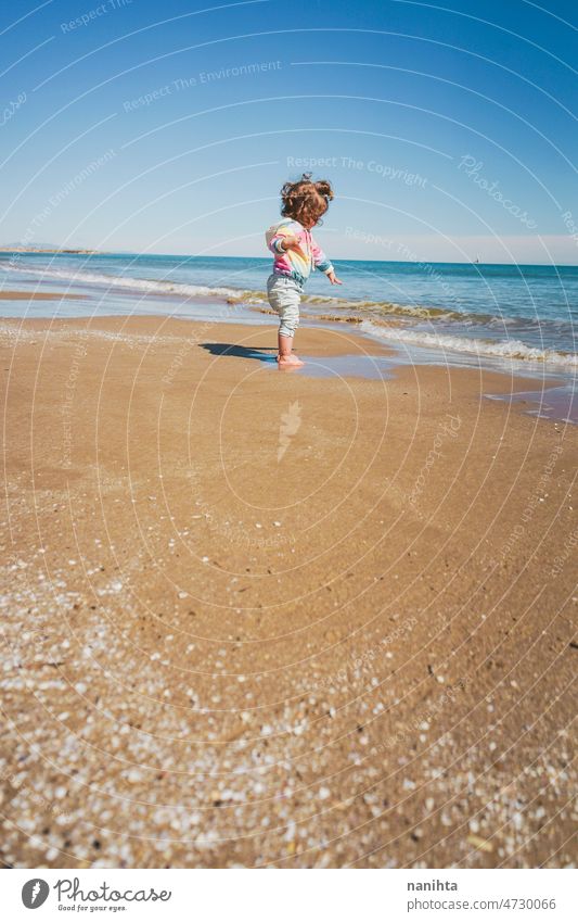 Wide view of a little girl wearing rainbow hoodie playing at the beach freedom happiness holidays baby toddler family sea shore playful real lifestyle people