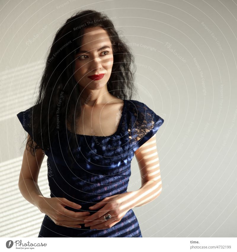 Woman in blue dress Feminine Dress Long-haired Dark-haired Stand Observe look View to the side Front view room sunny Strip of light