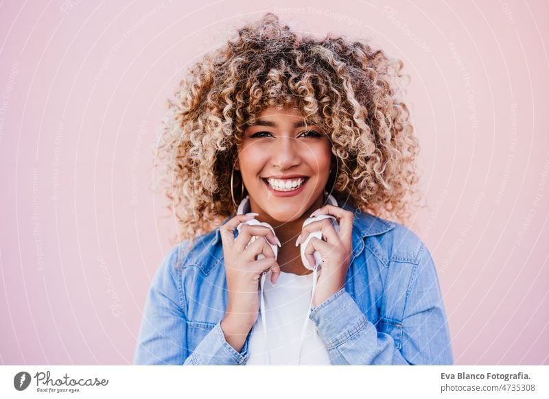 portrait of smiling hispanic woman with afro hair in city using mobile phone and headset. lifestyle listening body positivity curvy selfie technology internet