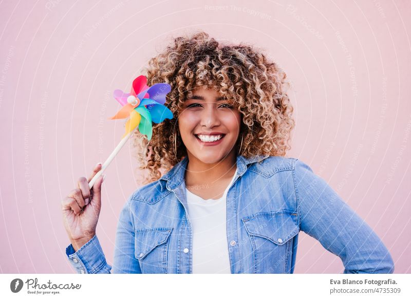 beautiful happy hispanic woman with afro hair holding colorful pinwheel. pink background,wind energy windmill sustainability reduce reused recycle city smiling