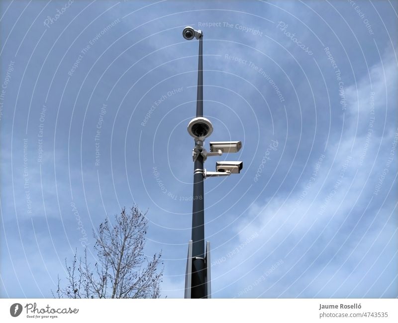 Mast with four surveillance cameras, three of them at the same height and the fourth at the top of the mast. In the background the blue sky with soft clouds and on one side you can see a tree from the top.