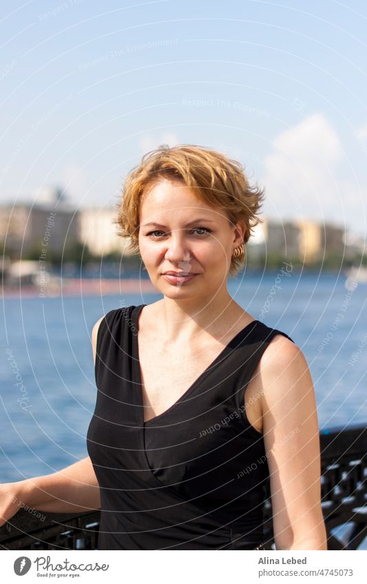 Portrait of a young girl with a short haircut. A girl on a sunny day poses on the embankment in a black dress. portrait of a girl blonde emotions attractive