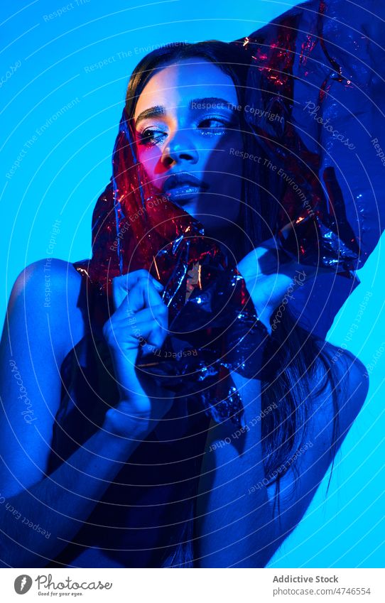 Fashionable woman wrapped in plastic in neon illumination model fashion provocative cool posture makeup female style blue light illuminate glow creative