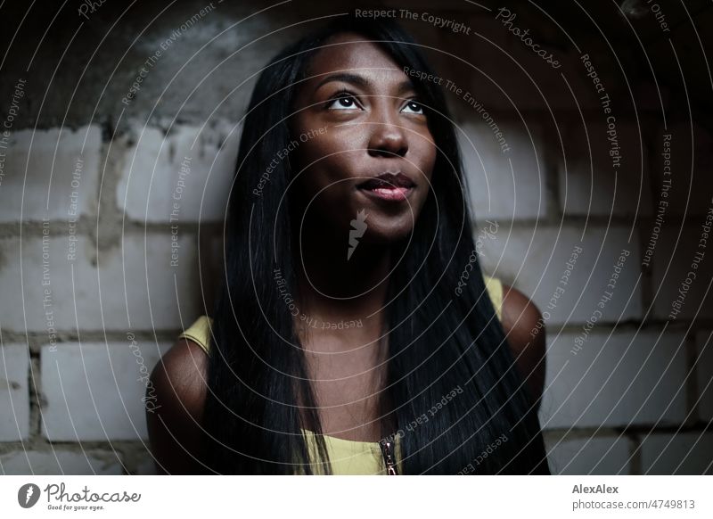 Young beautiful long haired woman in yellow summer dress stands in front of white brick wall in dark room and looks up to light Woman Young woman pretty