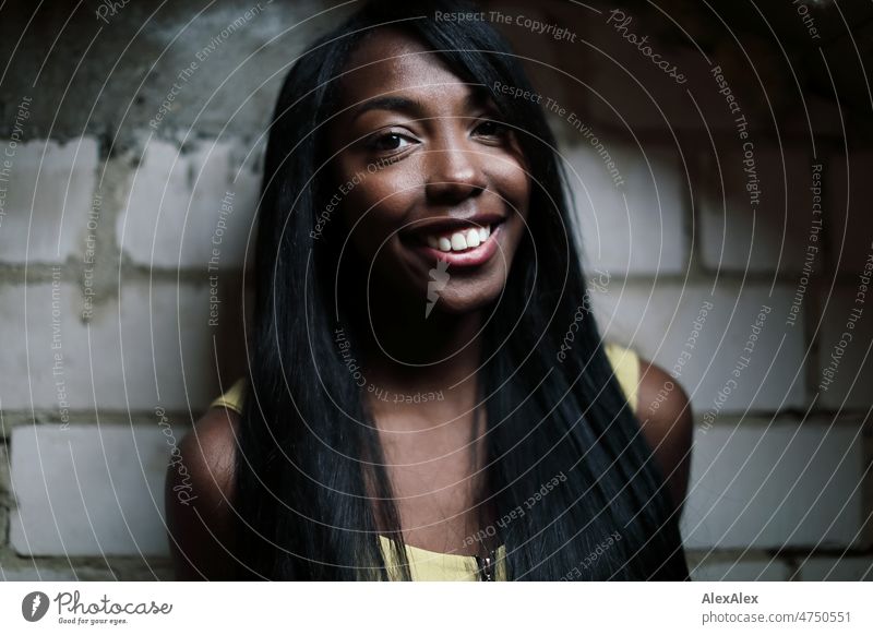 Young beautiful long haired woman in yellow summer dress stands in front of white brick wall in dark room and looks up to light Woman Young woman pretty