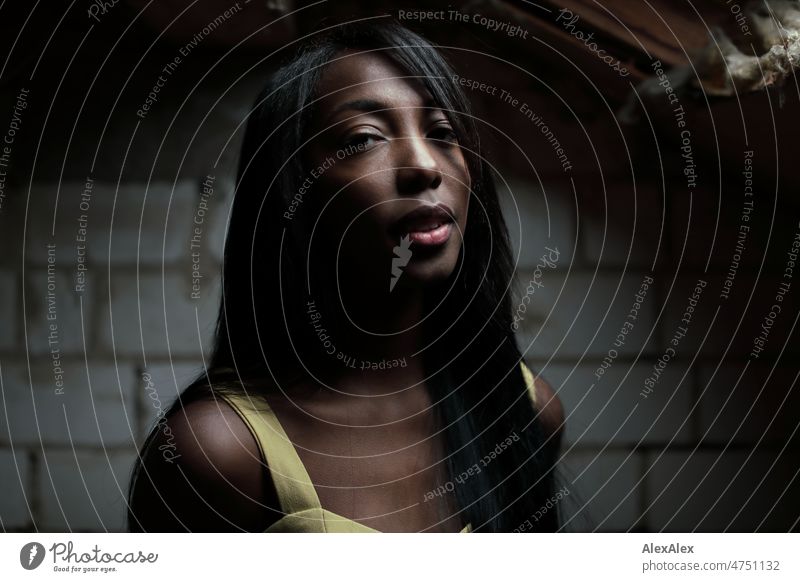 Young beautiful long haired woman in yellow summer dress stands in front of white brick wall in dark room and looks up to light Woman Young woman pretty