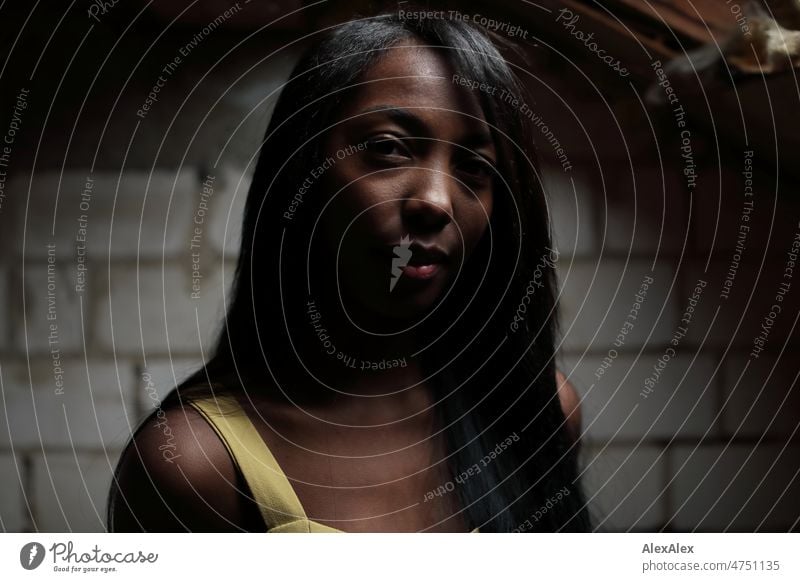 Young beautiful long haired woman in yellow summer dress stands in front of white brick wall in dark room and looks up to light Woman Young woman pretty