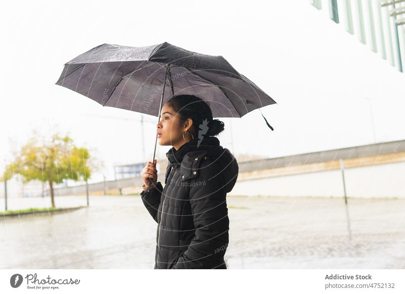 Ethnic woman with umbrella standing on street during rain overcast wow wonder reaction urban protect season female surprise jacket weather city mouth opened