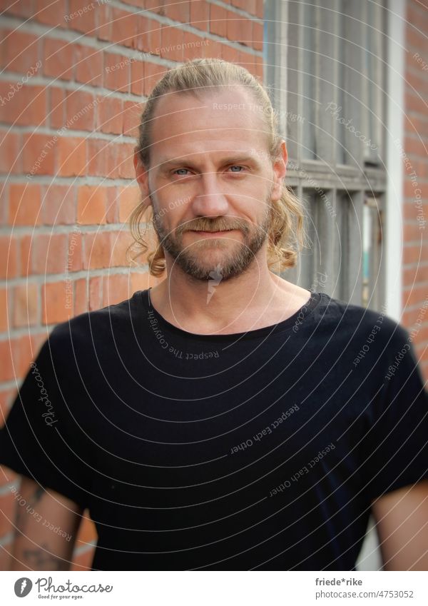 Man in front of brick wall portrait Facial hair Curl Brick Factory Black T-shirt Tattoo long hairs Blonde blue eyes smilingly Mysterious