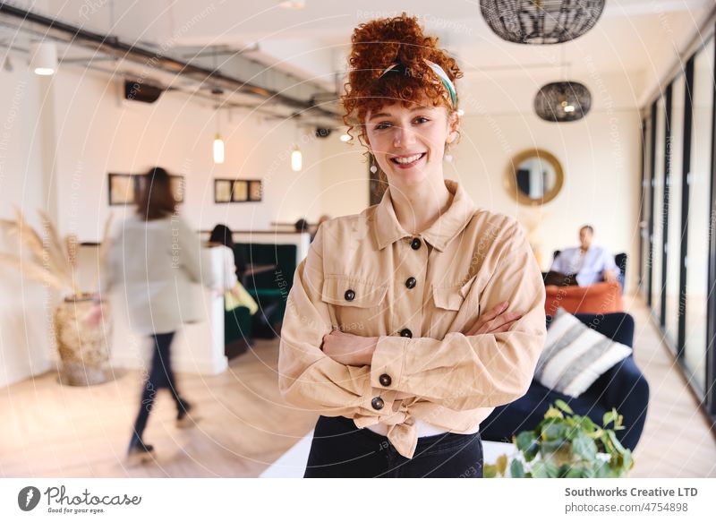 Portrait of confident young white woman with red curly hair smiling and looking at camera with arms folded in coworking space portrait female owned business